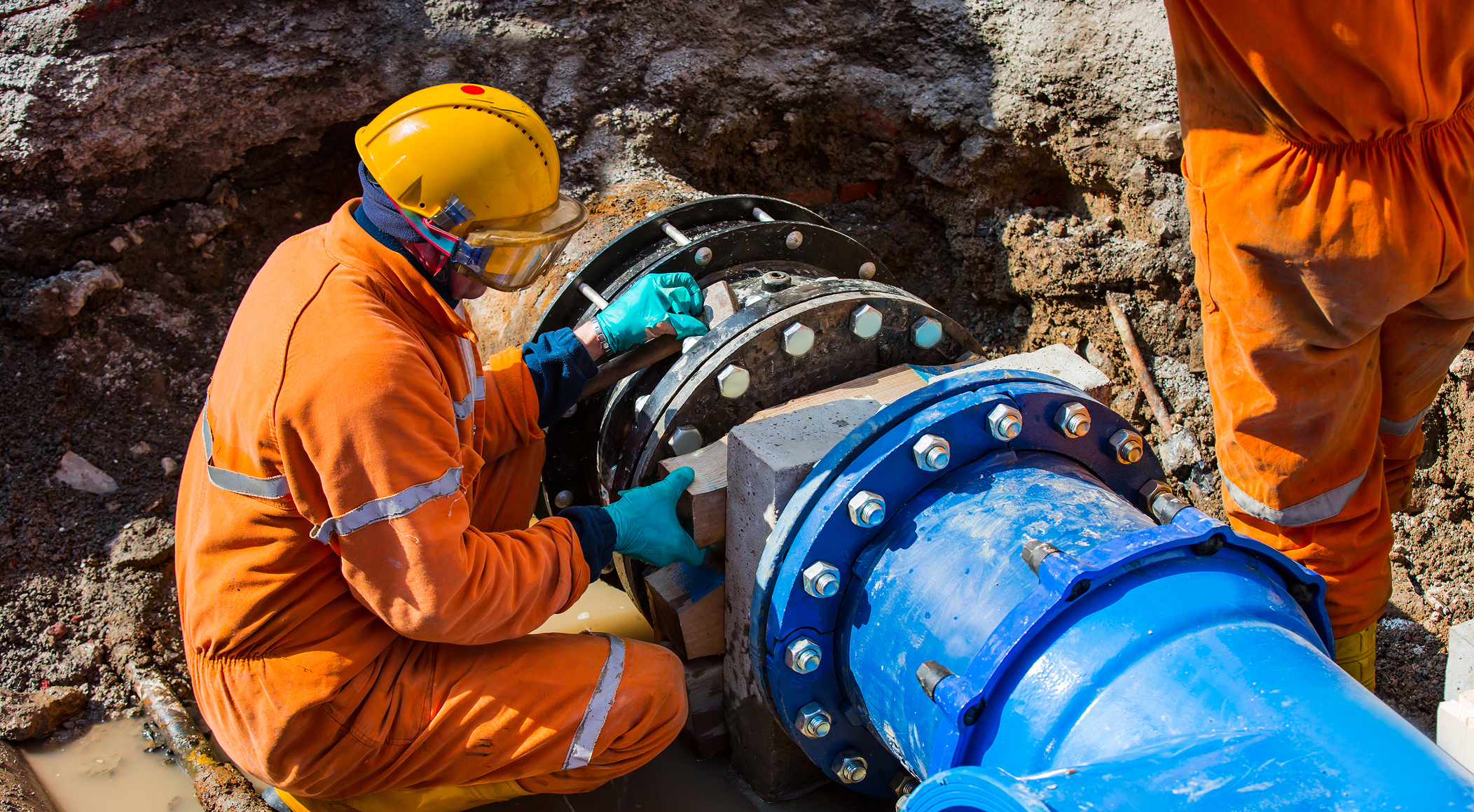 Browne Group - Worker in orange safety gear repairing a large blue industrial pipe at a muddy construction site.