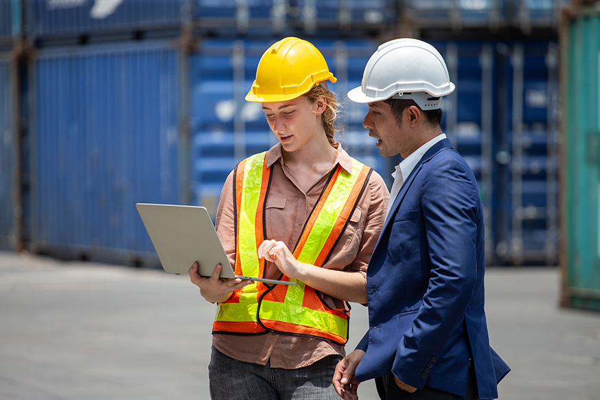 Browne Group - Two workers in safety helmets and vests look at a laptop together at an industrial worksite.