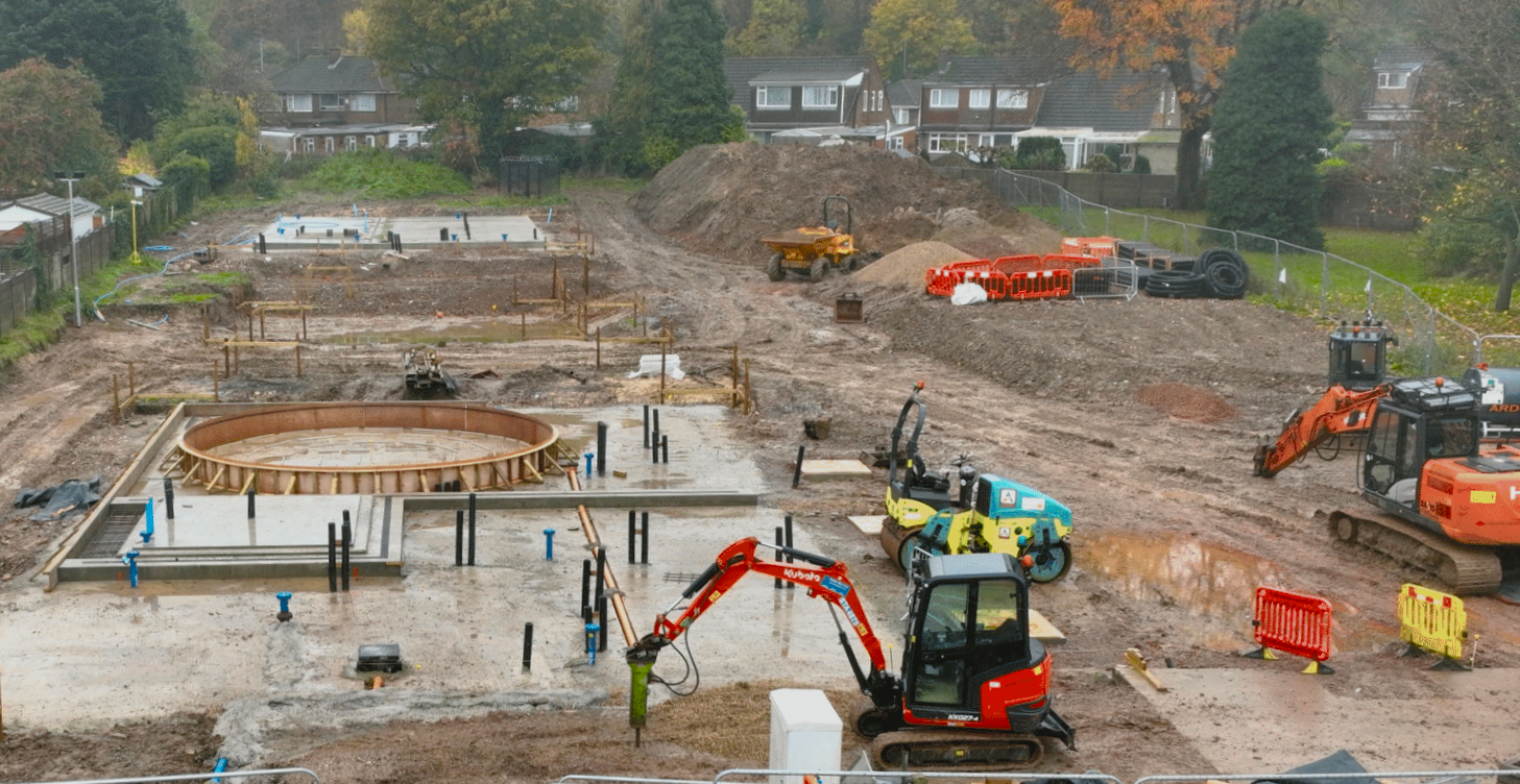 Browne Group - Construction site with excavators, concrete foundations, and building materials on muddy ground near houses.