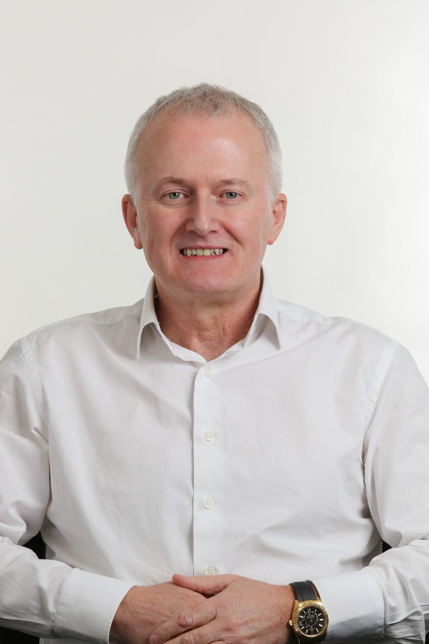 Browne Group - Man in a white dress shirt sitting and smiling with hands folded, against a plain light background.