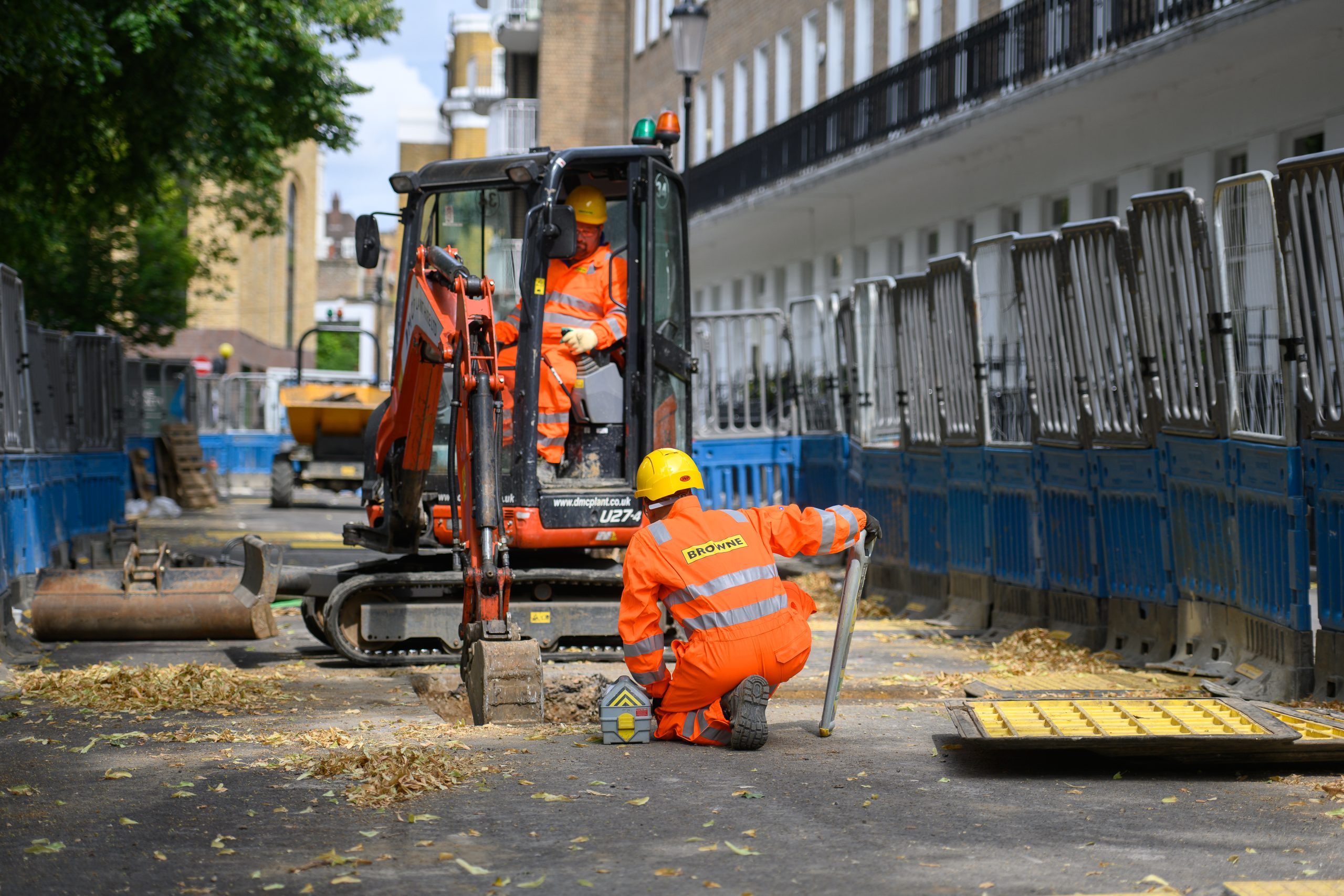 Browne Group - Two construction workers in orange gear operate machinery and work on a fenced-off urban street.