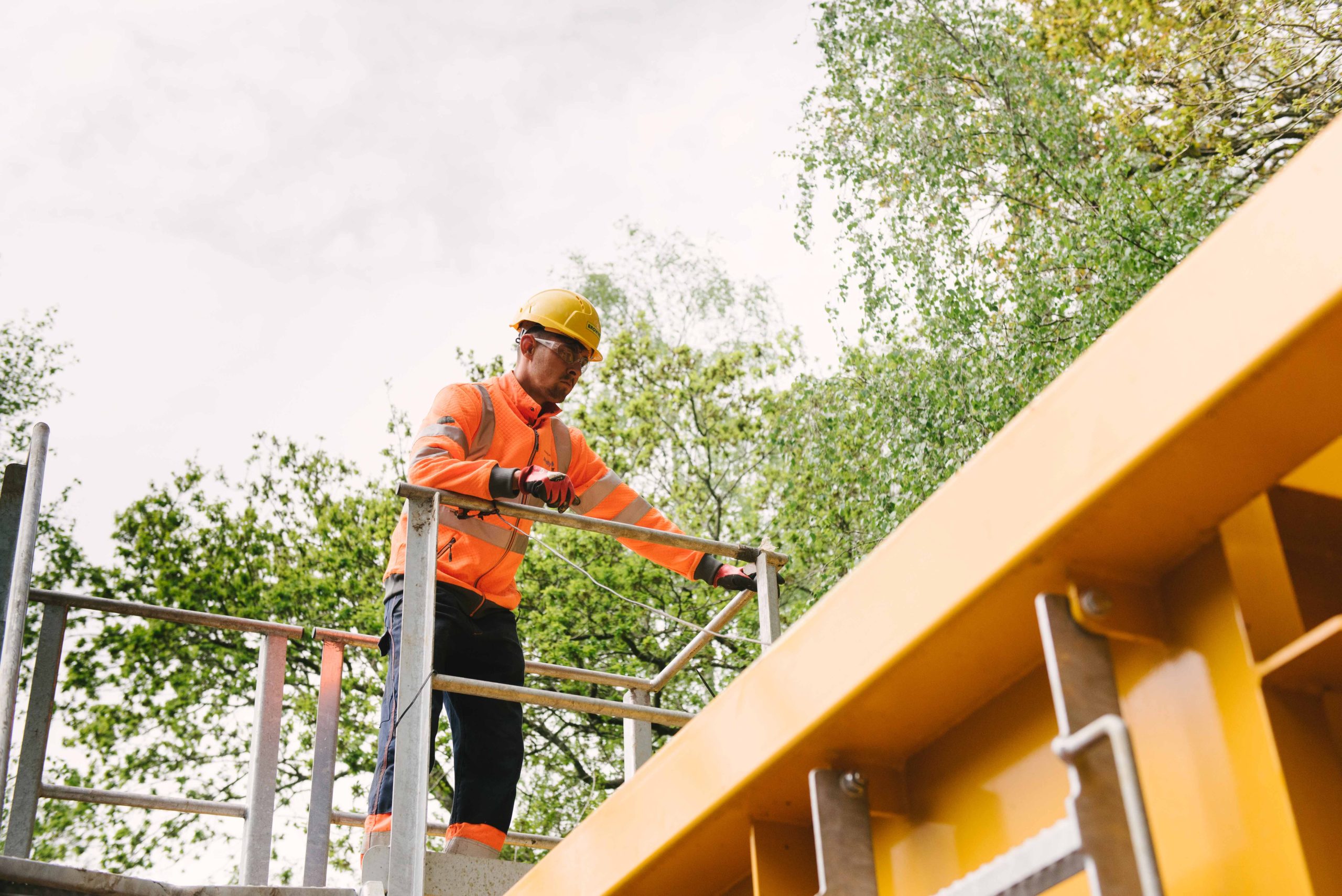 Browne Group - Construction worker in a hard hat and orange jacket stands on scaffolding outdoors near yellow equipment.