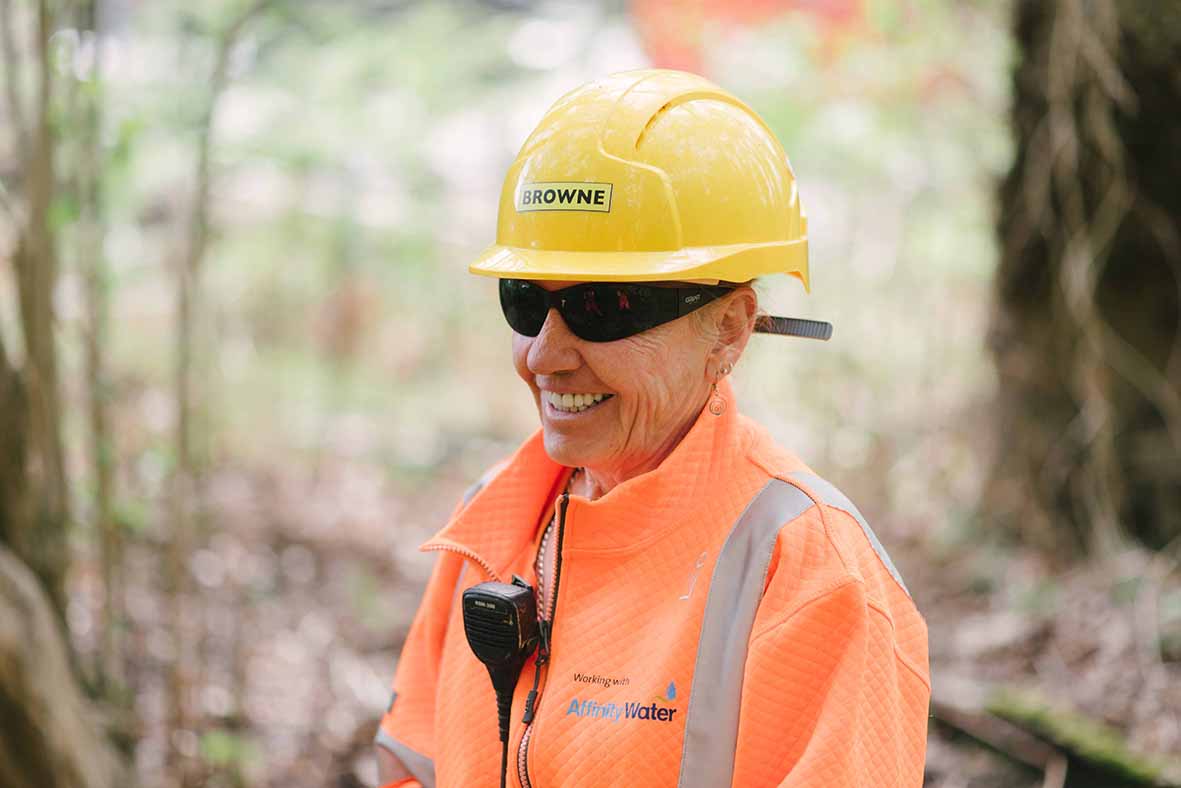 Browne Group - Smiling worker in an orange safety jacket and yellow hard hat stands outdoors, wearing sunglasses.