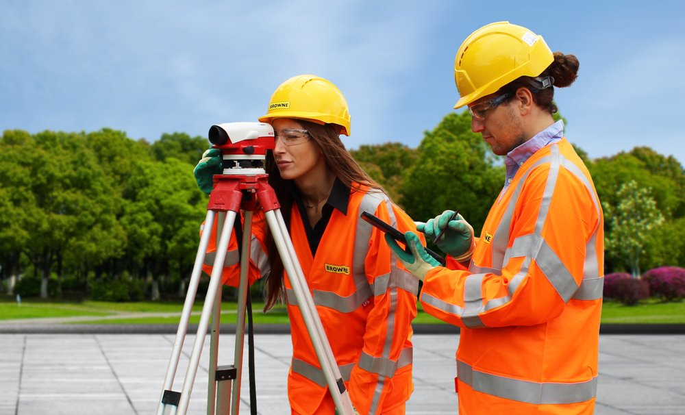 Browne Group - Two surveyors in orange safety gear use a leveling instrument outdoors on a sunny day.