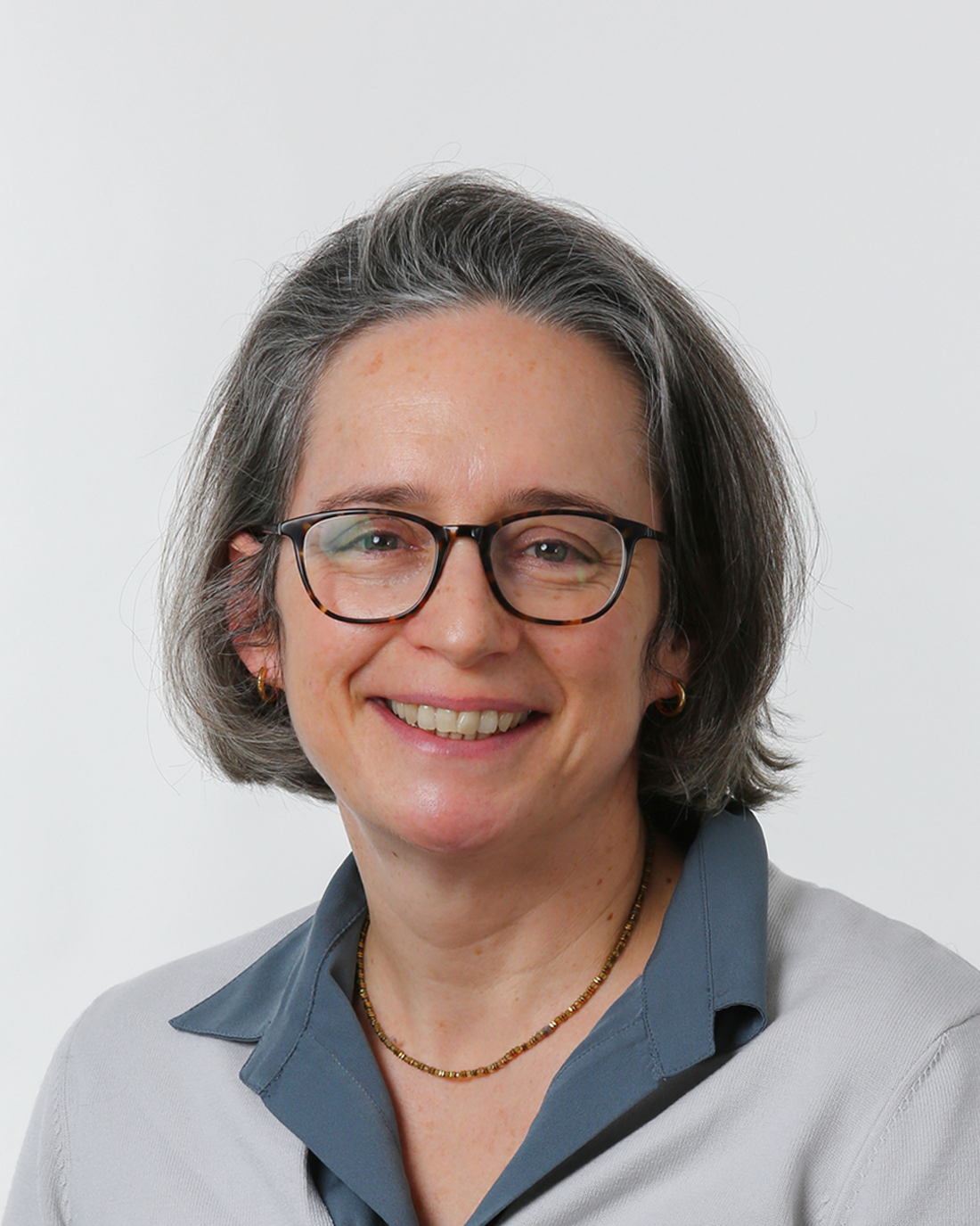Browne Group - A woman with short gray hair, glasses, and a blue collared shirt smiles at the camera against a white background.