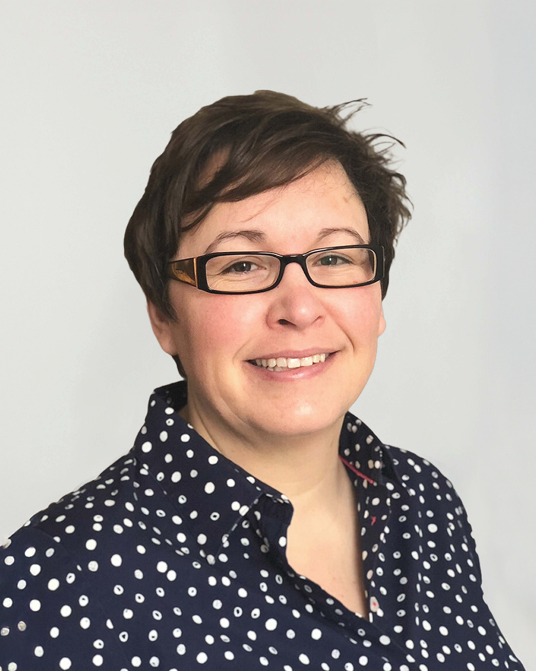 Browne Group - Smiling person with short brown hair, glasses, and a navy blue shirt with white polka dots.
