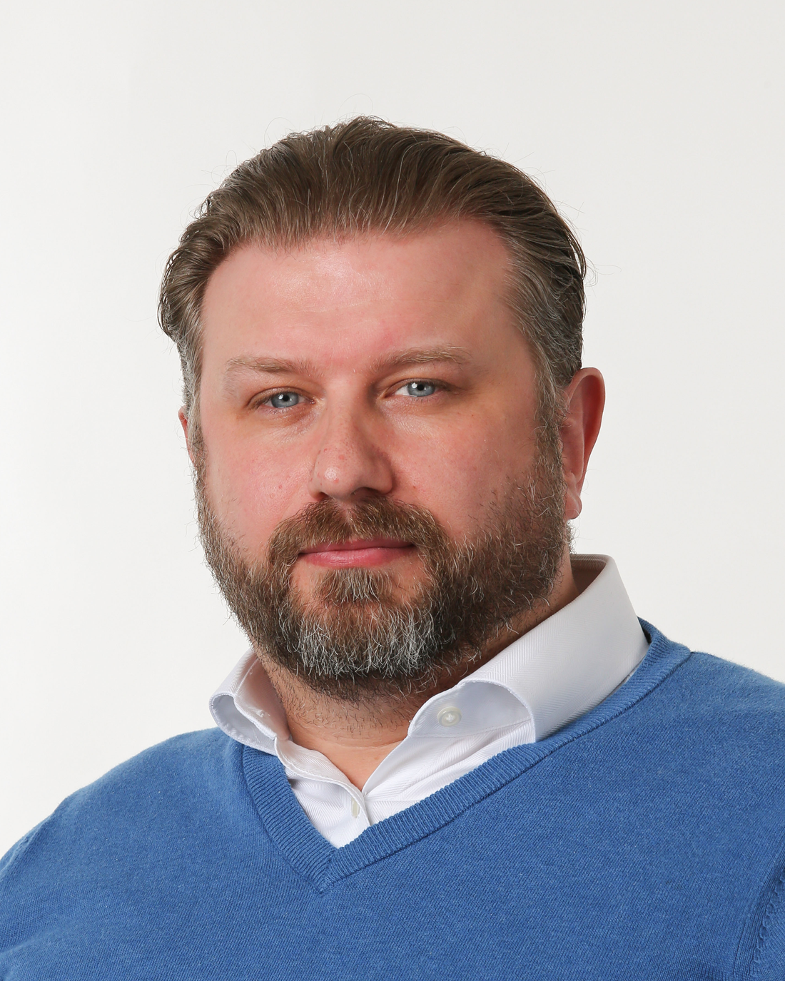 Browne Group - Man with short brown hair and beard wearing a blue sweater and white shirt, posing against a plain background.