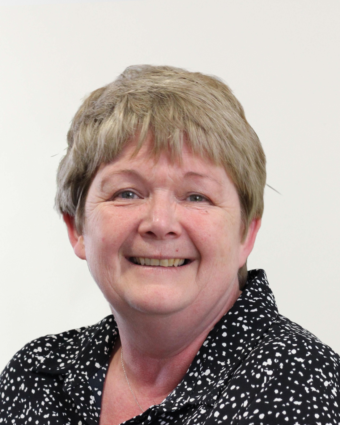 Browne Group - Smiling older woman with short light brown hair wearing a black and white patterned shirt on a plain background.